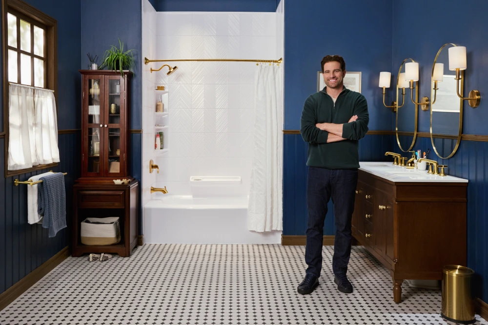 A smiling man stands with his arms crossed in a newly remodeled bathroom. The room features dark blue walls, a white and black tiled floor, and gold fixtures. The man is standing in front of a white bathtub and shower surround, and next to a wooden vanity with a white countertop and a mirror with gold light fixtures.